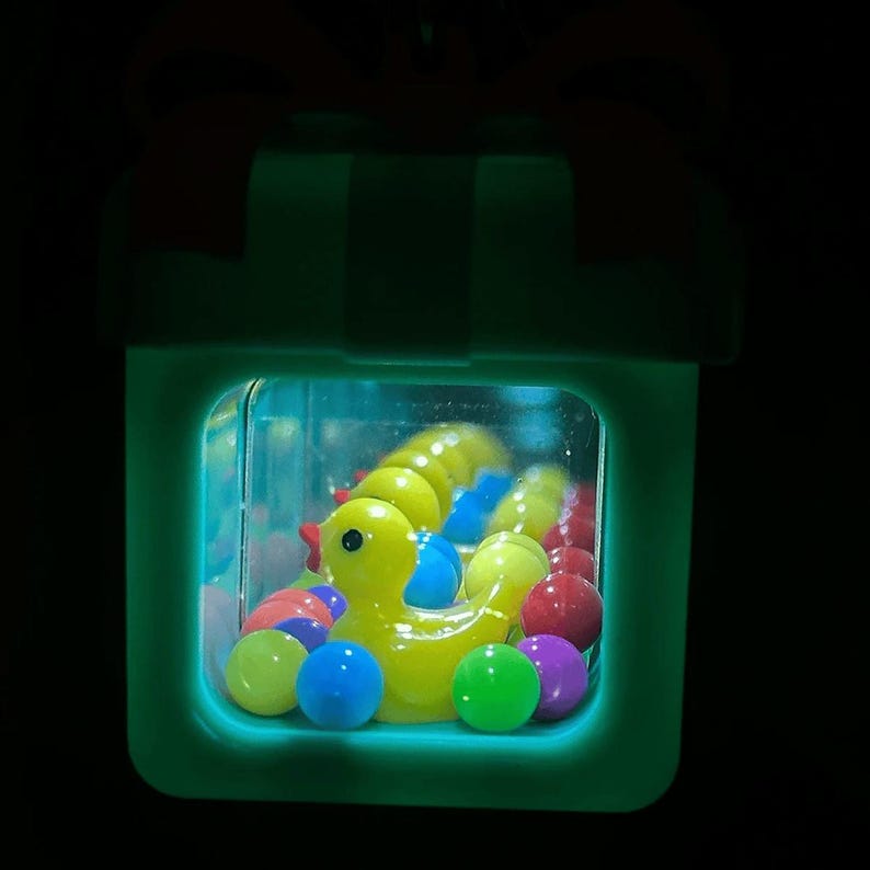 May include: A glowing, light-green, square-shaped container topped with a bow, illuminated from within. Inside, a bright yellow rubber duck is surrounded by numerous small, colorful, translucent spheres in shades of blue, red, and purple.
