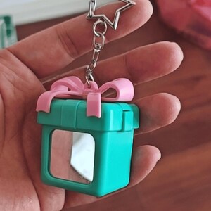 May include: A teal gift box shaped keychain featuring a light pink bow on top and a small square mirror embedded on the front face. The keychain includes a silver metal chain and a star-shaped clasp for attachment to keys or bags.