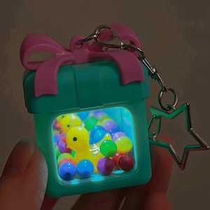 May include: A teal square keychain shaped like a gift box with a pink bow on top. The center is a transparent window containing a yellow rubber duck surrounded by small, colorful plastic spheres. The keychain includes a silver star-shaped charm.