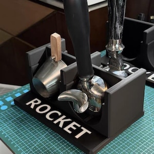 Puede incluir: Un soporte para accesorios de espresso impreso en 3D en color negro con la palabra ROCKET en letras blancas. El soporte sostiene un portafiltro de acero inoxidable con mango negro y un vaso dosificador de acero inoxidable con mango de madera clara.