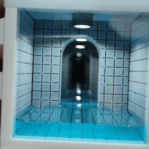 Könnte beinhalten: Eine Miniatur-Diorama-Box, die ein Schwimmbad mit hellblauen Fliesen an Wänden und Boden darstellt. Die Struktur enthält eine Reihe von Torbögen, die einen Tunneleffekt erzeugen und von kleinen Deckenleuchten beleuchtet werden, die sich im blauen Harzwasser spiegeln.
