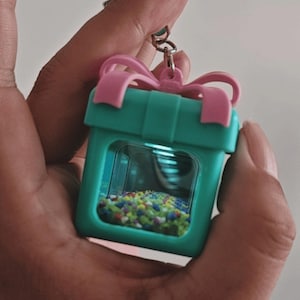 May include: A teal square-shaped keychain container designed to look like a gift box with a pink bow on top. The front features a clear window revealing a collection of small, multicolored beads inside the hollow center of the plastic structure.
