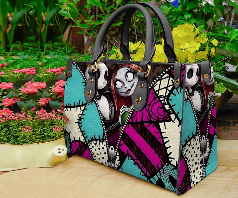 Puede incluir: Un bolso de mano estructurado con un estampado gr&aacute;fico de Jack Skellington y Sally de Pesadilla antes de Navidad. El dise&ntilde;o incluye un patr&oacute;n de parches en color turquesa, magenta y blanco con detalles de costuras negras y dos asas grises.