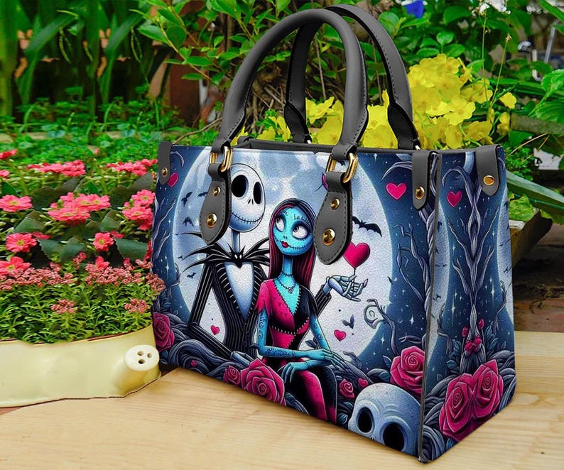 Op de afbeelding: Een rechthoekige handtas met een grafische print van een skeletfiguur en een blauwhuidig personage tegen een grote volle maan. Het ontwerp bevat roze rozen, kleine rode hartjes en donkere boomsilhouetten. Het heeft twee grijze hengsels met goudkleurige details.