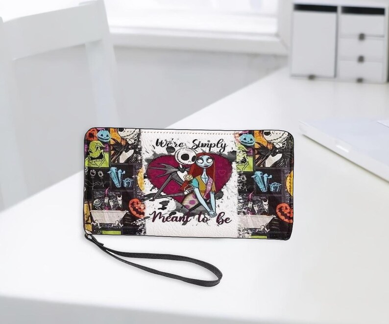 May include: A rectangular wallet with a wrist strap featuring a collage of characters from The Nightmare Before Christmas. The center displays Jack Skellington and Sally in front of a red heart with the text We're Simply Meant to be.