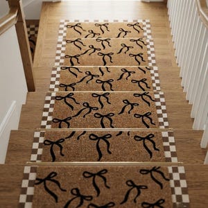 May include: A set of rectangular stair treads made of natural tan coir fiber, each featuring a repeating pattern of black bows. The treads are bordered by a white and tan checkered trim and are secured to wooden stairs with gold metal rods.