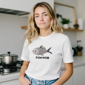 Può includere: Una t-shirt bianca a maniche corte con un'illustrazione grafica in bianco e nero di un pesce con un motivo testurizzato e il testo FISH MOB stampato in grassetto nero in lettere maiuscole sotto il grafico del pesce sul davanti.