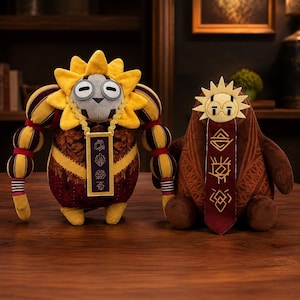May include: Two plush toys featuring stylised designs. The left figure has a yellow sun-shaped head, grey face, and maroon patterned body with a gold pendant. The right figure is brown with a sun-shaped head and a long maroon banner with symbols.