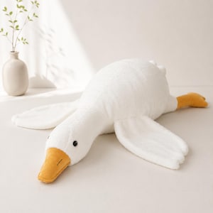 May include: A plush toy shaped like a white goose with a soft, textured fabric body, outstretched wings, and a bright yellow beak and feet. The goose has small, round black eyes and is positioned in a prone, resting posture on a flat surface.