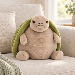 May include: A plush turtle toy with a soft, beige, rounded body and a textured, olive-green shell. The stuffed animal features a small head with dark eyes, a neutral expression, two short arms, and two short legs, all crafted from a fuzzy fabric.