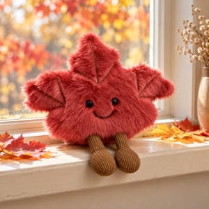 Op de afbeelding: Een pluche speeltje in de vorm van een rood esdoornblad met een pluizige textuur, voorzien van geborduurde zwarte ogen en een glimlachende mond. Het speelgoed heeft twee bungelende bruine ribfluwelen benen met ronde voeten op een vensterbank.