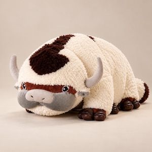 May include: A plush toy shaped like a six-legged bison with cream-coloured textured fur, a dark brown arrow-shaped marking on its head, grey horns, a grey muzzle, and dark brown hooves. The creature has large dark eyes and a soft, rounded body.