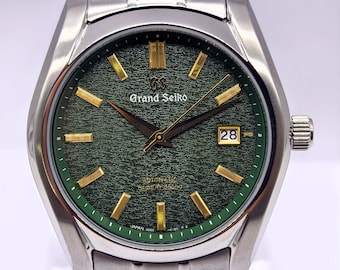Puede incluir: Un reloj automático Grand Seiko con esfera verde oscuro texturizada, marcadores de hora dorados y manecillas doradas. El reloj cuenta con una caja y brazalete de eslabones de acero inoxidable, una ventana de fecha a las tres y el texto Grand Seiko.