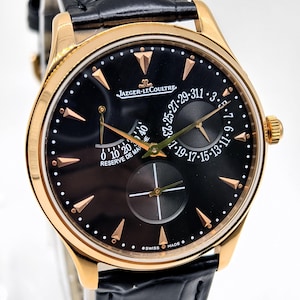 May include: A Jaeger-LeCoultre wristwatch with a round gold case, a black dial, and a black leather strap. The dial features gold-toned hour markers, a power reserve indicator labelled RESERVE DE MARCHE, a date sub-dial, and a small seconds sub-dial.