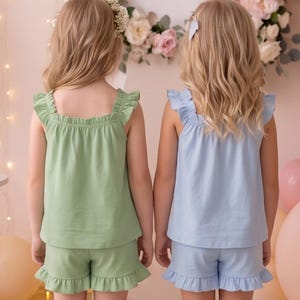 May include: Two sets of pajamas consisting of a sleeveless top and shorts with ruffled trim. One set is sage green and the other is light blue. Both tops feature a square neckline with gathered fabric and ruffled shoulder straps.