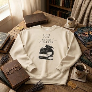 May include: A beige crewneck sweatshirt featuring a black graphic print of a dragon sleeping on a stack of books. The text above the dragon reads "JUST ONE more CHAPTER" in a mix of serif and script fonts, accented by small star symbols.