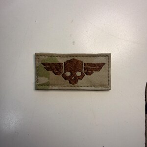 Puede incluir: Un parche rectangular de tela con un patr&oacute;n de camuflaje en tonos beige, arena y verde claro. Centrado en el parche hay un bordado de color marr&oacute;n oscuro que representa una calavera estilizada con alas extendidas horizontalmente a los lados.