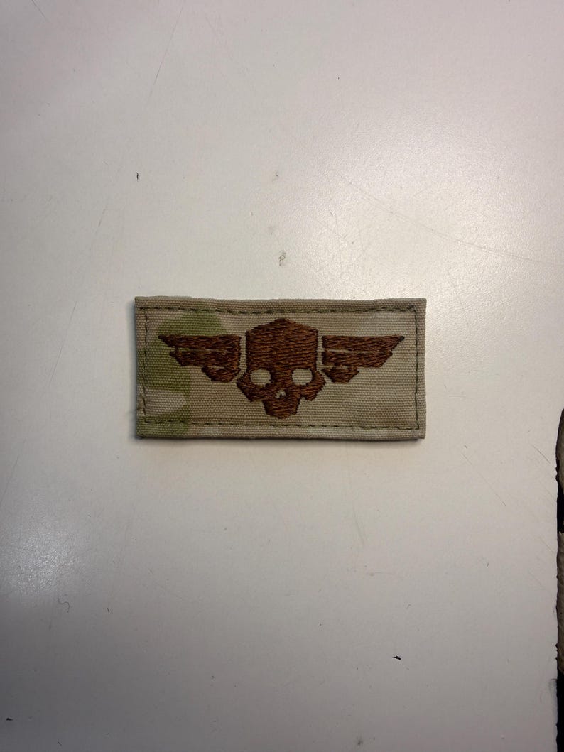 Puede incluir: Un parche rectangular de tela con un patr&oacute;n de camuflaje en tonos beige, arena y verde claro. Centrado en el parche hay un bordado de color marr&oacute;n oscuro que representa una calavera estilizada con alas extendidas horizontalmente a los lados.