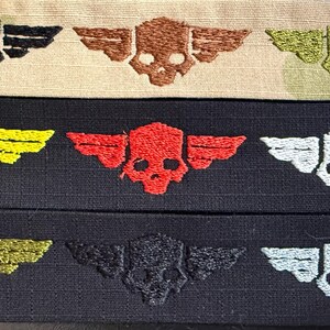 Puede incluir: Tres tiras de tela horizontales con parches bordados de calaveras aladas. La tira superior es beige con calaveras negras, marrones y verde oliva. La tira central es negra con calaveras amarillas, rojas y blancas. La tira inferior es negra con calaveras doradas, negras y blancas.