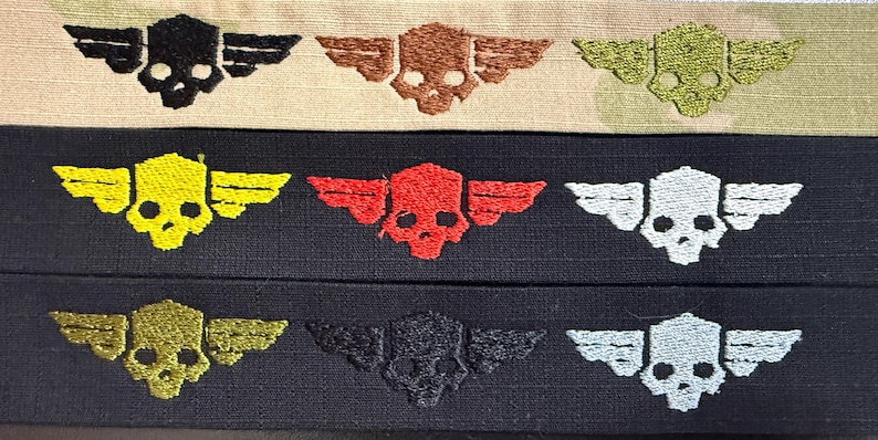 Puede incluir: Tres tiras de tela horizontales con parches bordados de calaveras aladas. La tira superior es beige con calaveras negras, marrones y verde oliva. La tira central es negra con calaveras amarillas, rojas y blancas. La tira inferior es negra con calaveras doradas, negras y blancas.