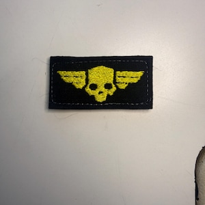 Puede incluir: Un parche rectangular negro que presenta una calavera amarilla bordada con alas estilizadas que se extienden desde los lados. El diseño está centrado en la tela, que tiene un borde cosido alrededor del perímetro de la forma rectangular.