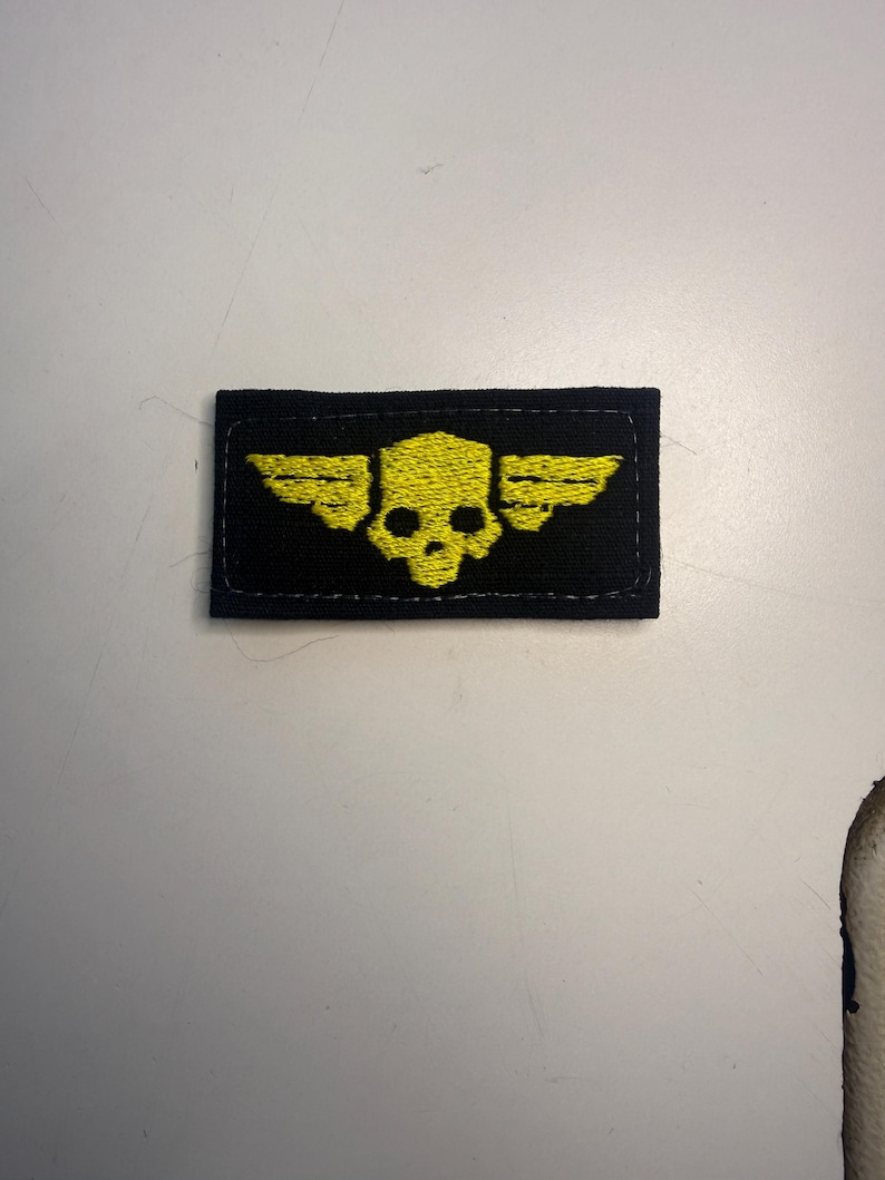 Puede incluir: Un parche rectangular negro que presenta una calavera amarilla bordada con alas estilizadas que se extienden desde los lados. El dise&ntilde;o est&aacute; centrado en la tela, que tiene un borde cosido alrededor del per&iacute;metro de la forma rectangular.
