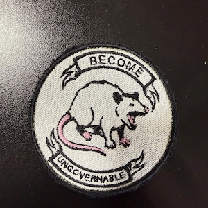 May include: A circular embroidered patch featuring a white opossum with pink accents on its tail and feet. The opossum is depicted with an open mouth. Above and below the animal are banners with the black text BECOME and UNGOVERNABLE.