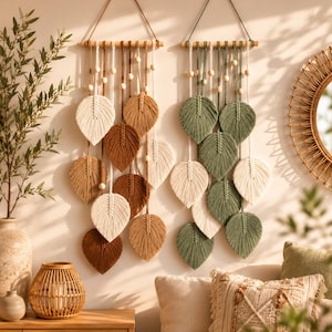 Op de afbeelding: Twee macramé wandhangers opgehangen aan houten stokken. Het linkerstuk bevat bladeren van crème, beige en bruin koord met houten kralen. Het rechterstuk toont bladeren van saliegroen en crème koord, eveneens versierd met houten kralen.