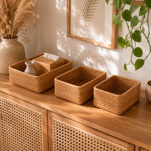 Pode incluir: Três cestos de armazenamento retangulares de vime trançado com um acabamento natural em castanho claro. Os cestos apresentam um padrão de trama em grelha consistente e apertado com topos abertos, dispostos em fila sobre uma superfície de madeira.
