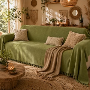 Puede incluir: Una funda de sofá texturizada en color verde salvia con un borde de flecos a juego en la parte inferior. El sofá está decorado con tres cojines beige con patrones tejidos intrincados y una manta beige drapeada con borlas.