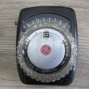 May include: A black and silver vintage light meter with a red GE logo. The meter has a dial with numbers 0-7 and a scale for frames per second. The meter also has a scale for time and a scale for light sensitivity.