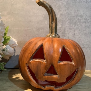 May include: A carved orange pumpkin with a smiling face, perfect for Halloween decorations. The pumpkin has triangular eyes and a wide, toothy grin. It features a long, curved stem and is set against a neutral background.