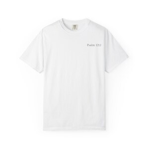 Op de afbeelding: Een wit t-shirt met korte mouwen, een ronde hals en de tekst Psalm 121:1 gedrukt in een klein, donker schreeflettertype op de rechterbovenkant van de borst. Het kledingstuk heeft een standaard pasvorm met een effen achterkant en een label.