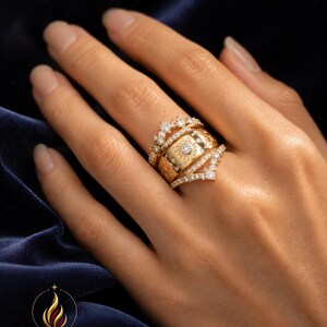 Könnte beinhalten: Ein Set aus drei Goldringen mit einem zentralen Band, das mit einem Sonnen- und Mondmotiv graviert ist, flankiert von zwei zarten Bändern mit kleinen klaren Steinen. Der mittlere Ring hat einen runden Stein, während die äußeren eine Kronenform bilden.