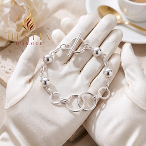 Op de afbeelding: Een zilverkleurige armband met een knevelsluiting, afgewisseld met gladde bolvormige kralen en een reeks in elkaar grijpende cirkelvormige schakels. Het sieraad is voorzien van de tekst HESTIA D'AMOUR en een vlamlogo linksboven.