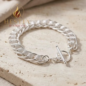 May include: A silver chain bracelet featuring a series of textured, spherical links alternating with smooth, interlocking oval links. The bracelet is secured with a toggle clasp consisting of a circular ring and a straight bar closure.