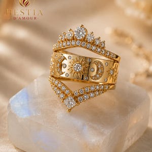 May include: A gold ring set consisting of three stacked bands. The top band is a crown shape with small stones, the middle band features engraved sun and moon motifs with inset stones, and the bottom band is a V-shaped chevron with stones.