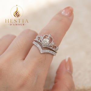 May include: A silver-toned ring set featuring a crown-shaped band with a central round stone and a V-shaped pavé band. The design includes intricate metalwork and small clear stones. The text HESTIA D'AMOUR is visible in the upper left corner.