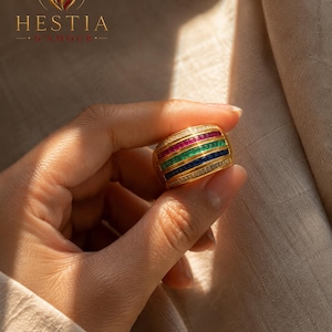 Puede incluir: Un anillo de banda ancha de oro con tres filas horizontales de piedras preciosas de corte baguette engastadas en canal en rojo, verde y azul, bordeadas por dos filas exteriores de pequeñas piedras claras engastadas en pavé sobre tela.