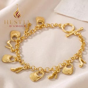 Puede incluir: Una pulsera de dijes dorada con una cadena de eslabones circulares que sostiene colgantes en forma de bolsos y zapatos de tacón alto. La pulsera se asegura con un cierre de barra y tiene el texto HESTIA D'AMOUR impreso en la parte superior.