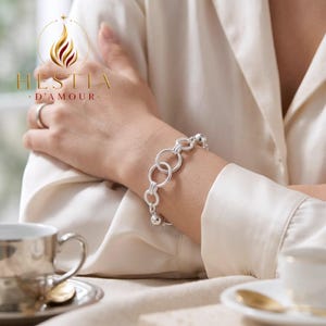 May include: A silver chain bracelet featuring a series of interlocking circular links and small spherical beads. The bracelet is worn on a wrist, with the HESTIA D'AMOUR logo and text visible in the upper left corner of the frame.