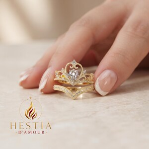 Pode incluir: Um conjunto de anéis de ouro com uma banda em forma de coroa com uma pedra central em forma de pera e uma banda em V a condizer, ambas incrustadas com pequenas pedras claras. O logótipo HESTIA D'AMOUR é visível no canto inferior esquerdo.