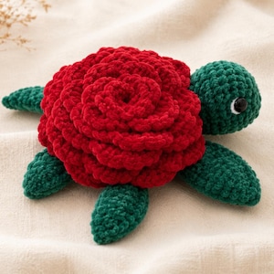 Op de afbeelding: Een gehaakt schildpadspeeltje met een donkergroene kop, vier donkergroene flippers en een schild opgebouwd uit gelaagde, dieprode garenblaadjes die lijken op een bloeiende roos. De schildpad heeft één zichtbaar zwart-wit plastic oog.