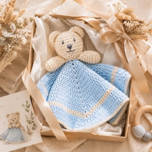 May include: A crochet baby security blanket featuring a light beige teddy bear head attached to a light blue and cream-colored square blanket with a textured stitch pattern, presented in a gift box with a matching illustrated card and wooden ring.