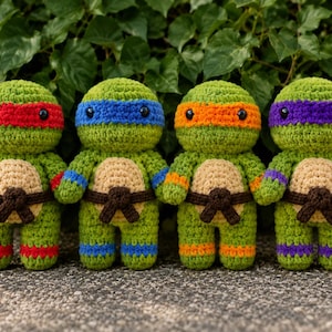 Op de afbeelding: Vier gehaakte schildpaddenpoppen met groene lichamen en beige buiken. Elke pop draagt een bruine riem en een gekleurd masker en polsbandjes: rood, blauw, oranje en paars. Ze hebben zwarte plastic ogen en staan in een rij naast elkaar.