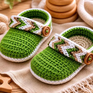 Op de afbeelding: Een paar handgehaakte babyschoentjes in een levendige groene kleur. Elke schoen heeft een bandje met een wit en bruin chevronpatroon over de bovenkant, vastgezet met een ronde houten knoop aan de zijkant. De zolen en randen zijn wit.