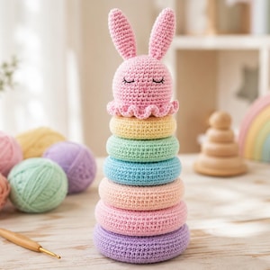 Pode incluir: Um brinquedo de empilhar em crochê com uma cabeça de coelho rosa no topo, com olhos fechados e orelhas longas, sobre um eixo vertical. O brinquedo inclui seis anéis texturizados nas cores amarelo, verde-menta, azul-claro, pêssego, rosa e roxo.