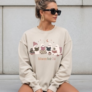 Könnte beinhalten: Ein beigefarbenes Sweatshirt mit Rundhalsausschnitt und einer Grafik von vier geisterhaften Figuren, die Bücher lesen. Unter den Geistern steht der Text "Halloween-Book-Club", umgeben von kleinen Sternen, einer Mondsichel, Büchern und einer Kerze.