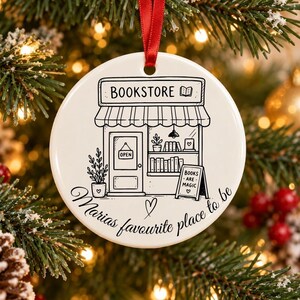 Könnte beinhalten: Ein rundes, weißes Keramikornament mit rotem Band, das eine schwarze Strichzeichnung eines Buchladens mit offenem Schild, Bücherregalen und einem Schild mit der Aufschrift BOOKS ARE MAGIC zeigt. Darunter steht Marias favourite place to be.