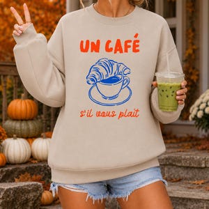 Könnte beinhalten: Ein beigefarbenes Sweatshirt mit Rundhalsausschnitt und einem Grafikdruck einer blauen Kaffeetasse mit einem darauf liegenden Croissant. Der Text "UN CAFÉ" steht in Orange darüber und "s'il vous plaît" in Orange darunter.